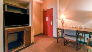 Ein Hotelzimmer mit Fernseher, einer roten Tür, einem Tisch mit Lampe und einem gemusterten Teppich.