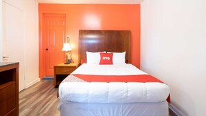 A hotel room with a bed, a nightstand with a lamp, a wooden headboard, and a door. - Premium King Bed | Jonesboro (Jonesboro)