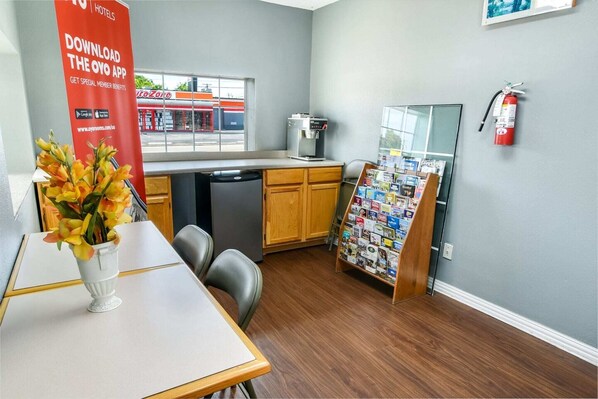A hotel room with a kitchenette, a dining area with a table and chairs, a vase with flowers, a fire extinguisher, and a bulletin board with pamphlets.