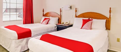 Habitación de hotel con dos camas, cada una con una colcha roja y blanca, cabecero de madera, mesita de noche con lámpara y ventana con cortina roja.