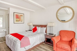 Habitación de hotel con cama, una silla, mesita de noche, lámpara, espejo y un cuadro en la pared.