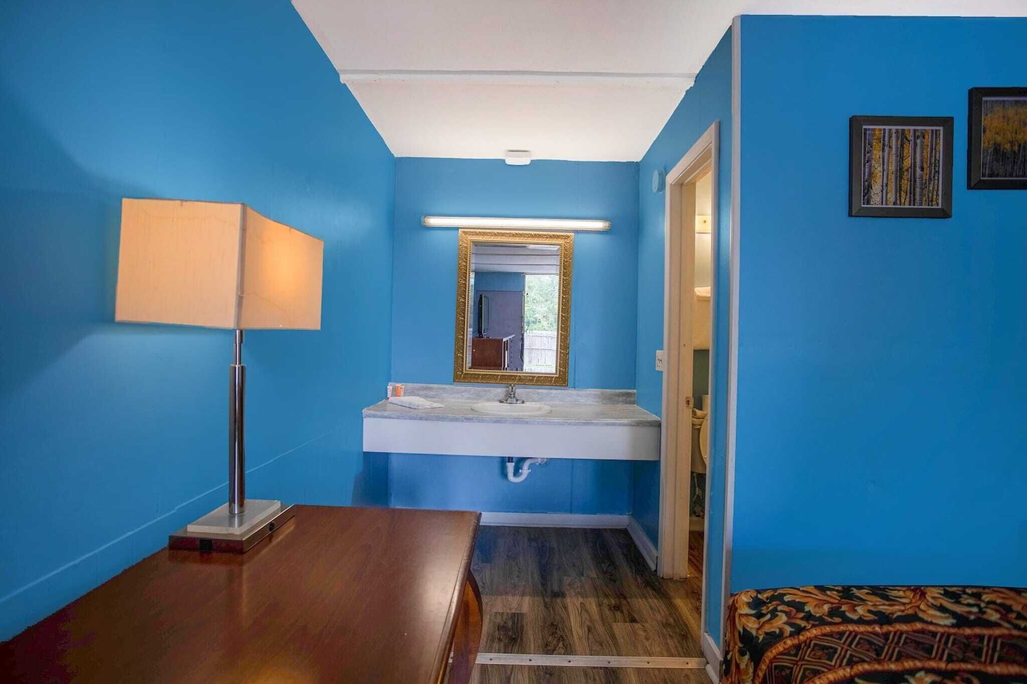 Uma casa de banho com parede azul, pia branca, espelho com moldura dourada, chão de madeira e tapete estampado.