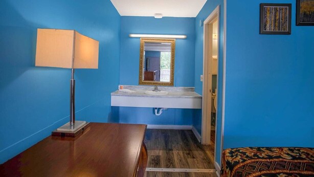 Un baño con pared azul, lavamanos blanco, espejo con marco dorado, piso de madera y alfombra estampada.