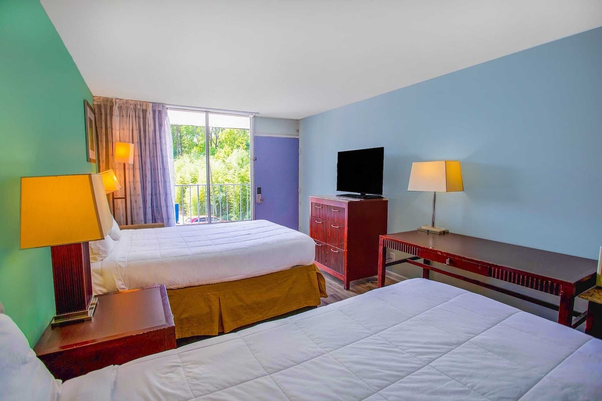 Ein Hotelzimmer mit zwei Betten, einem Fernseher, einem Schreibtisch und einem Balkon mit Blick auf Grünflächen.