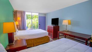 Ein Hotelzimmer mit zwei Betten, einem Fernseher, einem Schreibtisch und einem Balkon mit Blick auf Grünflächen.