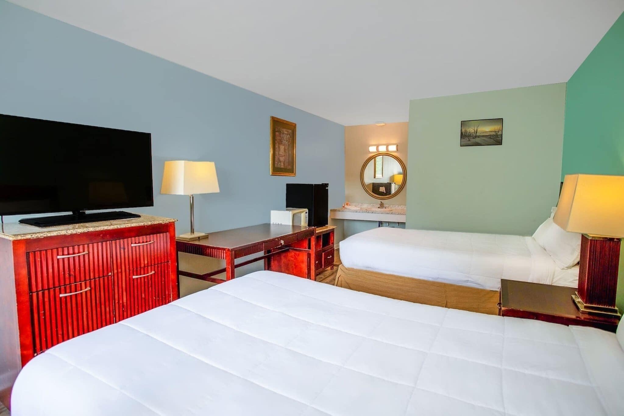 Quarto de hotel com duas camas, televisão, escrivaninha, espelho e luminária.