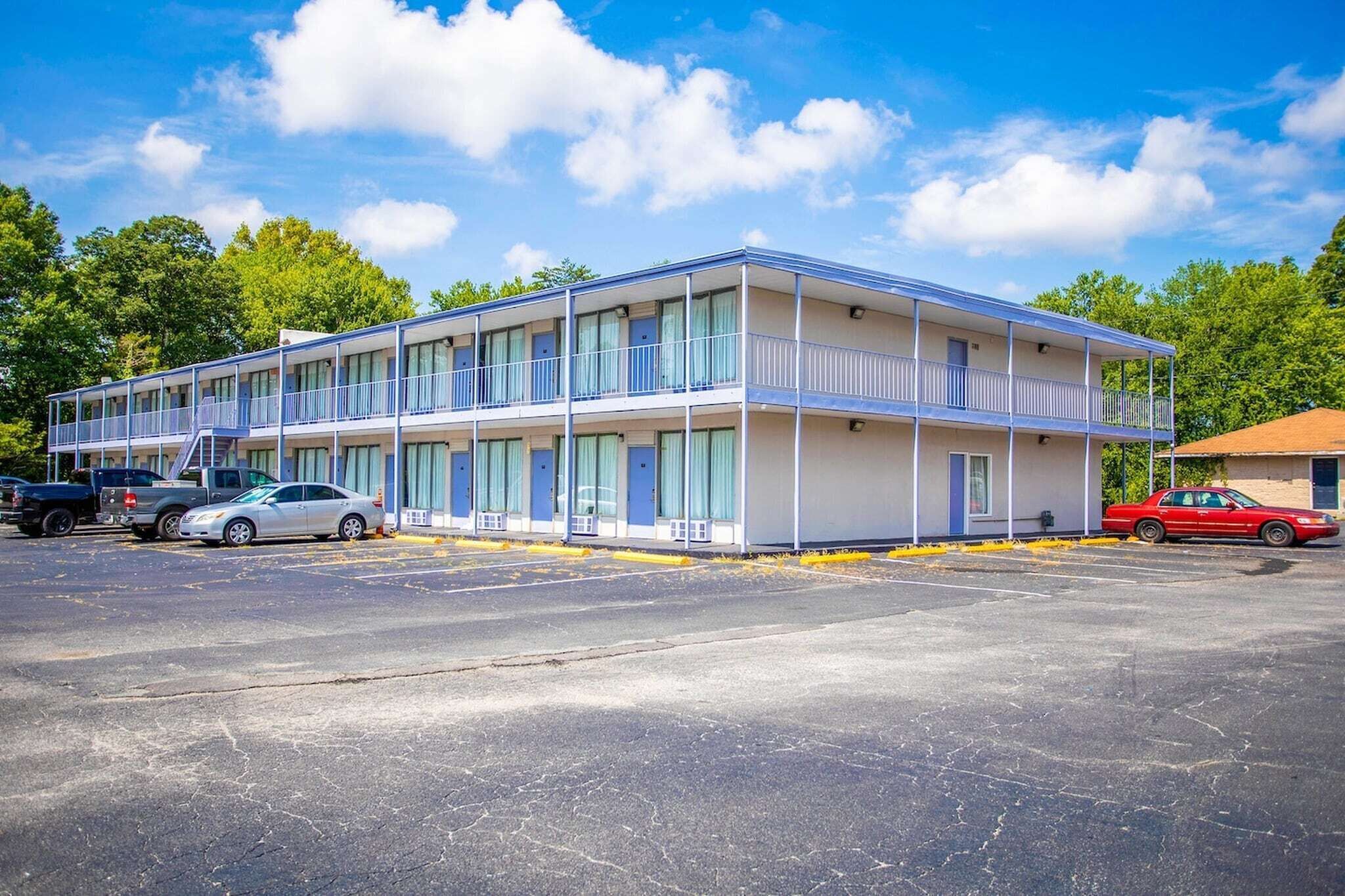 Un motel composé d’un bâtiment à deux étages, d’un espace de stationnement couvert et d’une voiture rouge garée devant.