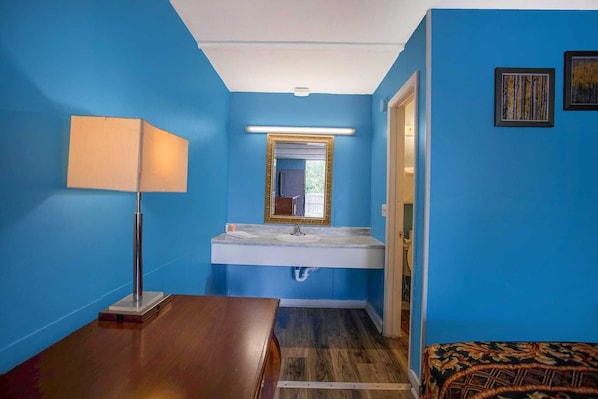 A bathroom with a blue wall, a white sink, a mirror with a gold frame, a wooden floor, and a patterned rug.