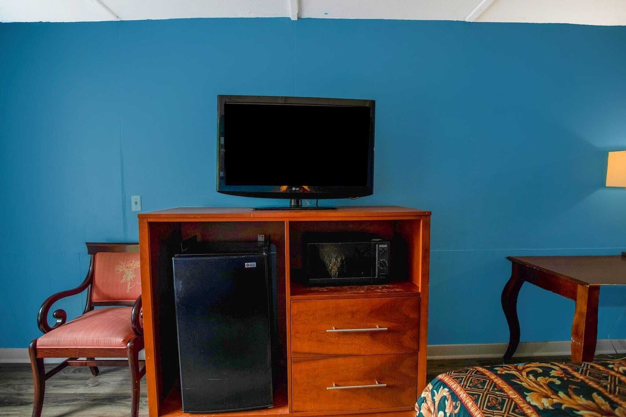 Une pièce comprenant un téléviseur posé sur un meuble en bois, un réfrigérateur, un four à micro-ondes, une chaise et une petite table.
