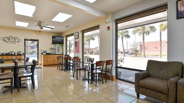 Un vestíbulo de hotel con zona de comedor, televisor y un amplio ventanal con vista a palmeras.