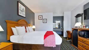 A hotel room with a bed, bedside table, lamp, dresser, microwave, and a mirror. - Lake Park GA | King Bed (Lake Park GA)