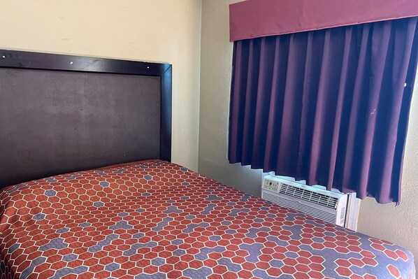 Ein Bett mit gemustertem Überwurf, ein dunkles Kopfteil, ein Fenster mit violetten Vorhängen und ein Klimagerät.