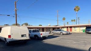 A parking lot with a white van, a white pickup truck, and a silver sedan parked in front of a motel with a red awning and palm trees in the background.