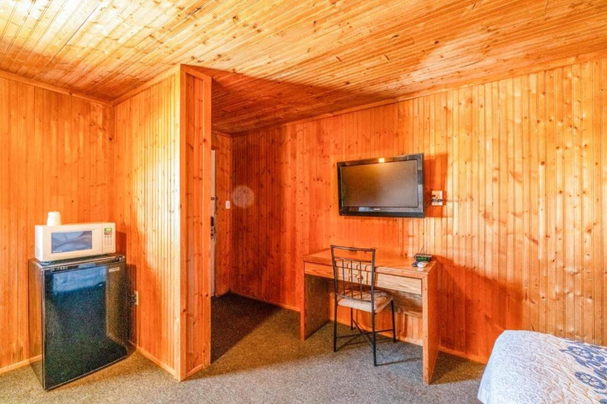 Ein Zimmer mit Holzwänden, einem Bett, einem Schreibtisch, einem Sessel, einem Fernseher und einem kleinen Kühlschrank.
