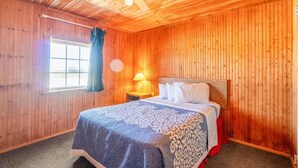 A wooden-paneled room with a bed, a ceiling fan, a window with curtains, and a nightstand with a lamp.
