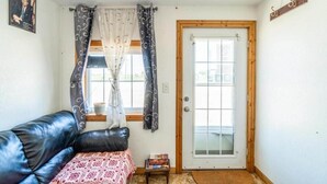 Una habitación con un sofá de cuero negro, una mesita con libros, una puerta con panel de vidrio y una ventana con cortinas.