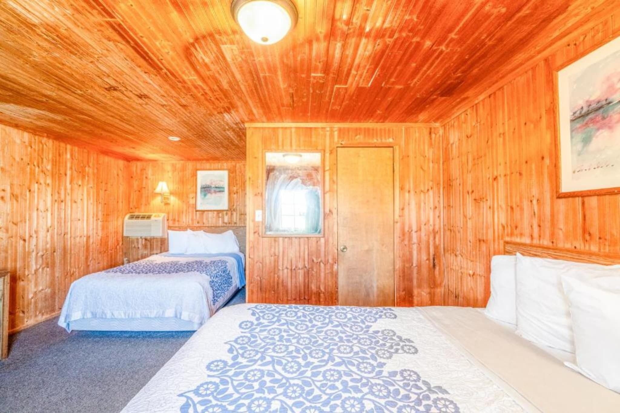 A wooden room with a bed, a door, a wall-mounted lamp, and a framed picture.