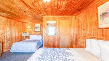 A wooden room with a bed, a door, a wall-mounted lamp, and a framed picture.