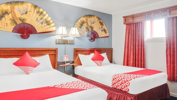 Een hotelkamer met twee bedden, rood-witte beddengoed, een houten hoofdbord, een ventilator, een lamp en een raam met rode gordijnen.