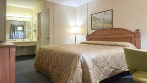 Una habitación de hotel con cama, escritorio, silla, espejo, lámpara y un cuadro en la pared.