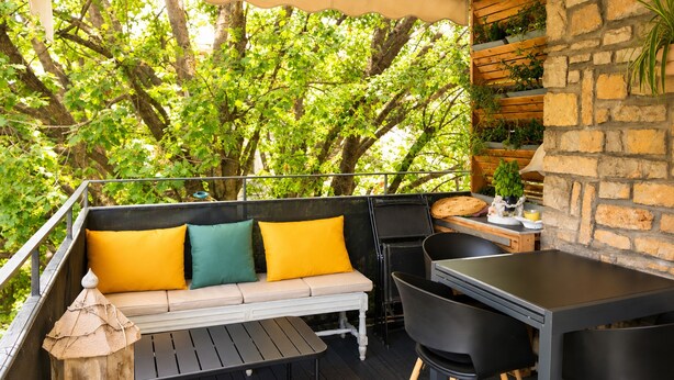 An outdoor balcony with a wooden table, black chairs, a sofa with yellow cushions, and a stone wall with greenery. - 225 Le Club C in Cannes (Cannes)