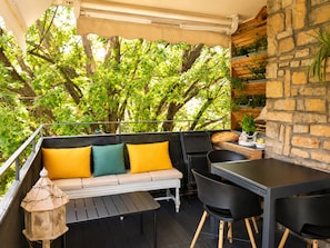 An outdoor balcony with a wooden table, black chairs, a sofa with yellow cushions, and a stone wall with greenery. - 225 Le Club C in Cannes (Cannes)