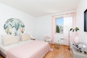 Ein Schlafzimmer mit Bett, Stuhl, einem Fenster mit Vorhängen, einem kleinen Tisch und einer Pflanze.