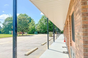 A covered outdoor area with a brick building, a paved parking lot, and trees in the background.