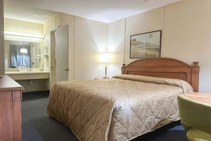 A hotel room with a bed, a desk, a chair, a lamp, a mirror, and a picture on the wall.