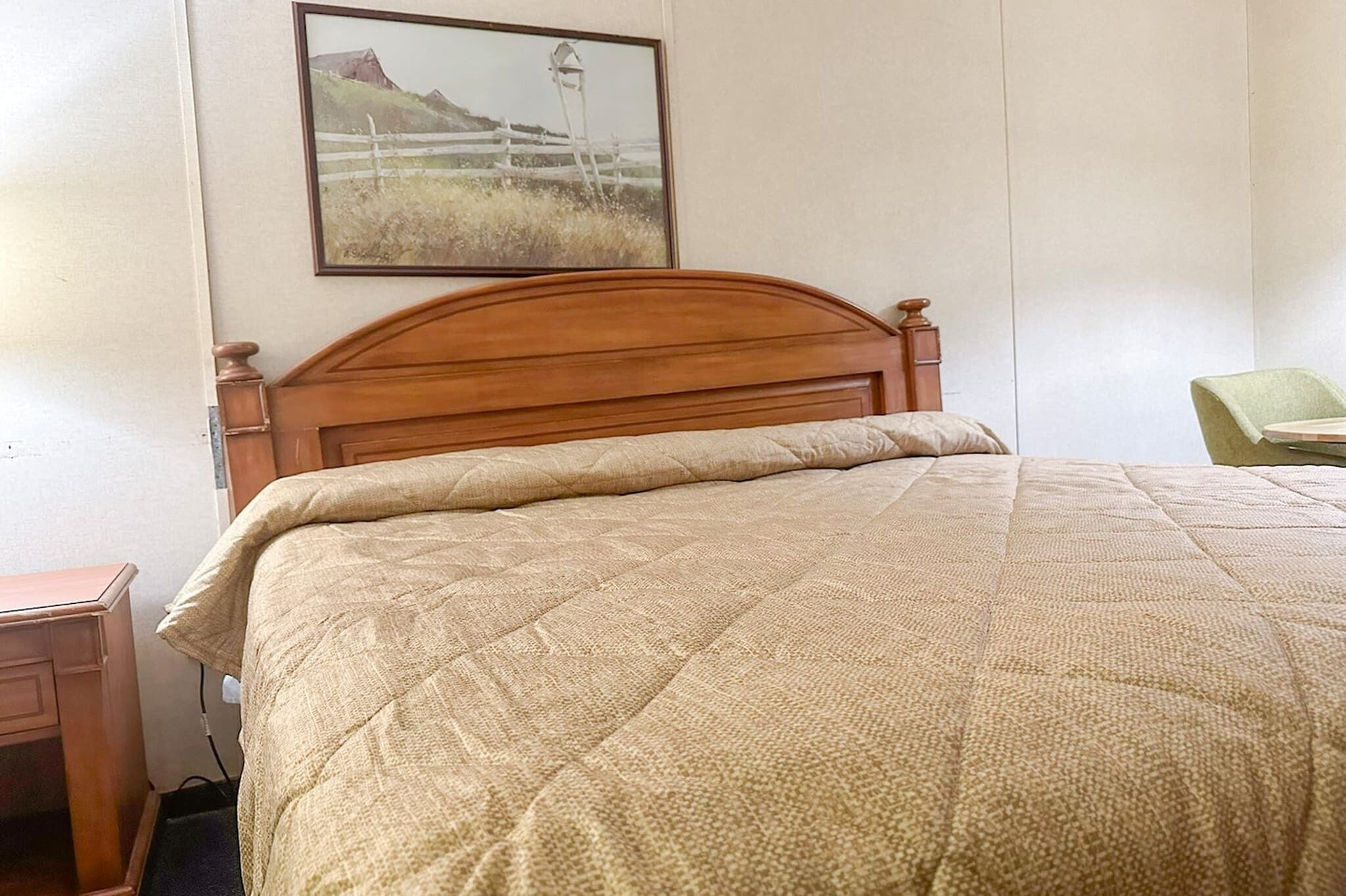 A bedroom with a wooden headboard, a beige quilted bedspread, a nightstand, and a framed picture on the wall.