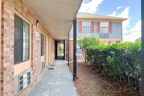 A covered walkway leading to a building with brick walls and a window with a red shutter.