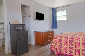 Ein Zimmer mit einer Mikrowelle, einem Holzkleiderschrank, einem Bett mit einem gemusterten Bettbezug, einem an der Wand montierten Fernseher und einem Fenster mit blauen Vorhängen.