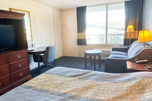 A hotel room with a bed, a desk, a chair, a TV, a lamp, and a view of the outside. - King Bed with Kitchenette Non-Smoking (Milledgeville, GA)