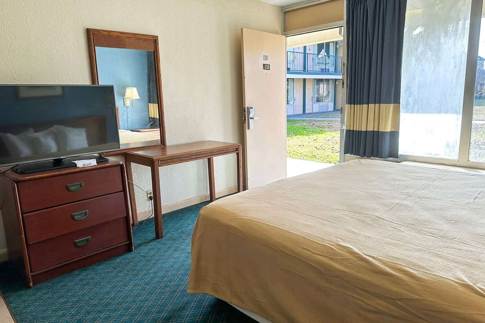 A hotel room with a bed, a TV on a wooden dresser, a desk, and a door leading to an outdoor area.