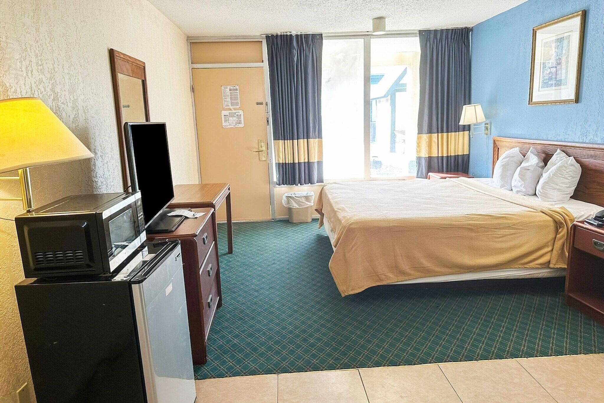 Una habitación de hotel con una cama, un escritorio con microondas, una cómoda, un cesto de basura y una ventana con cortinas.