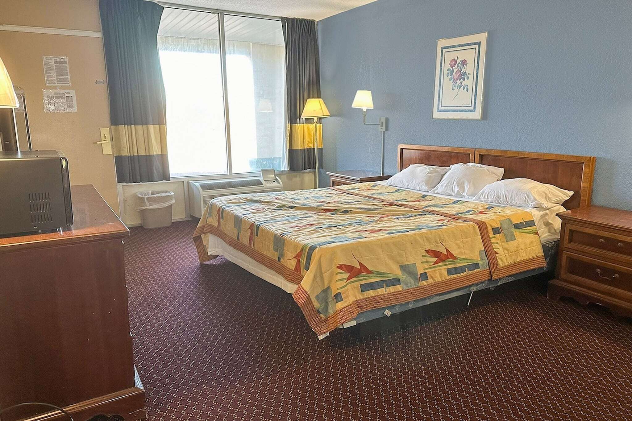 Et hotelværelse med seng, skrivebord, lampe, et billede på væggen og et vindue med gardiner.