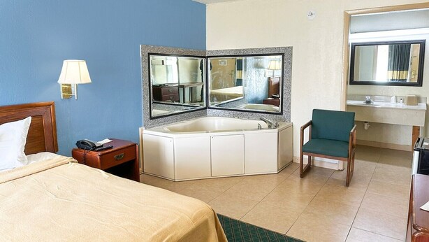 Een hotelkamer met een bed, een hoekbad, een stoel, een bureau, een spiegel en een nachtkastje.