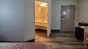 Een hotelkamer met een houten vloer, een bed, een bureau met stoel, een badkamer met wastafel en handdoekhouder, en een deur met slot en bordje.
