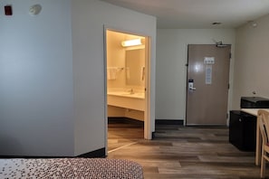 Habitación de hotel con piso de madera, una cama, un escritorio con silla, baño con lavamanos y perchero, y una puerta con cerradura y cartel.