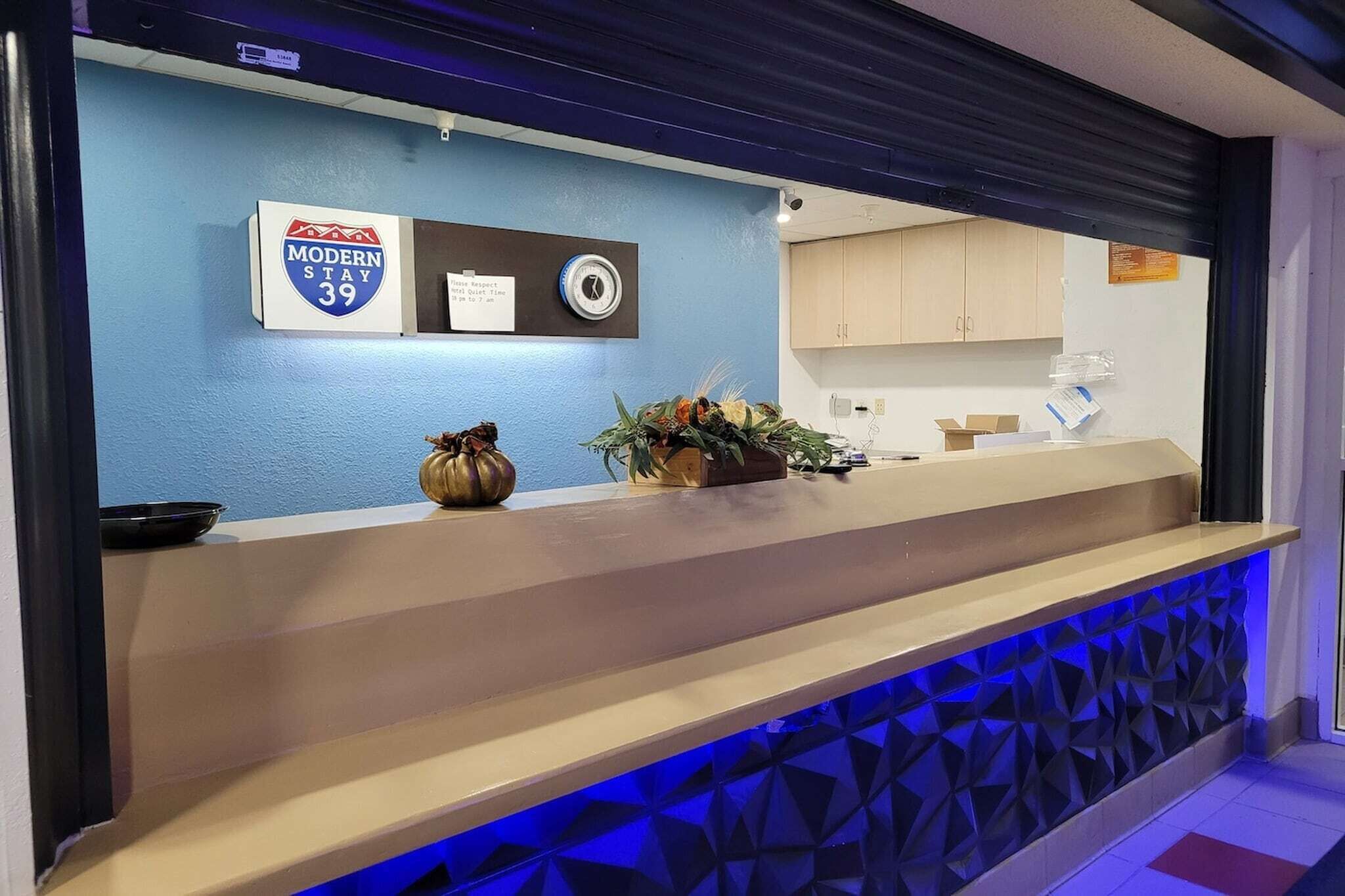 Un comptoir d’accueil avec un mur bleu, une enseigne indiquant « Modern Stay 39 », une horloge et une plante.