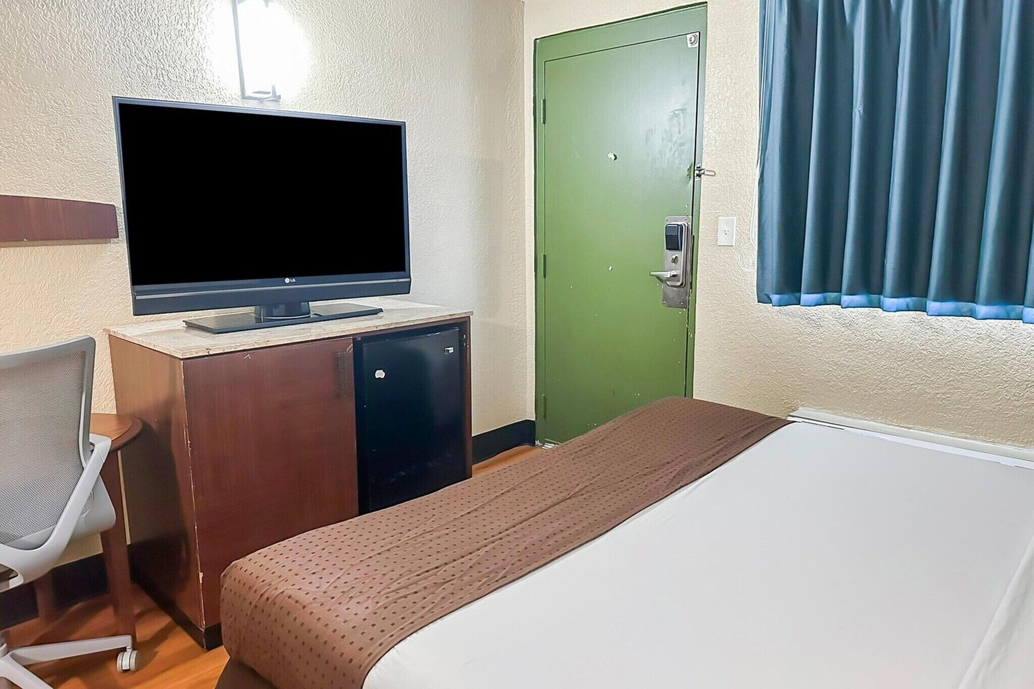 Quarto de hotel com uma cama, uma mesa de trabalho com televisão, uma porta verde e uma janela com cortinas.
