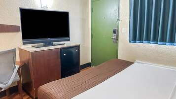 Quarto de hotel com uma cama, uma mesa de trabalho com televisão, uma porta verde e uma janela com cortinas.
