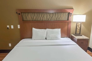 Hotellihuone, jossa on puinen sängynpääty, kaksi valkoista tyynyä, valkoinen peitto, yöpöytä lampun kanssa ja puinen kaappi.