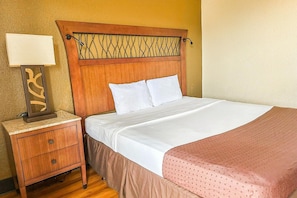 Habitación de hotel con cabecera de madera, lámpara de noche, cama con ropa blanca y almohada decorativa rosa.