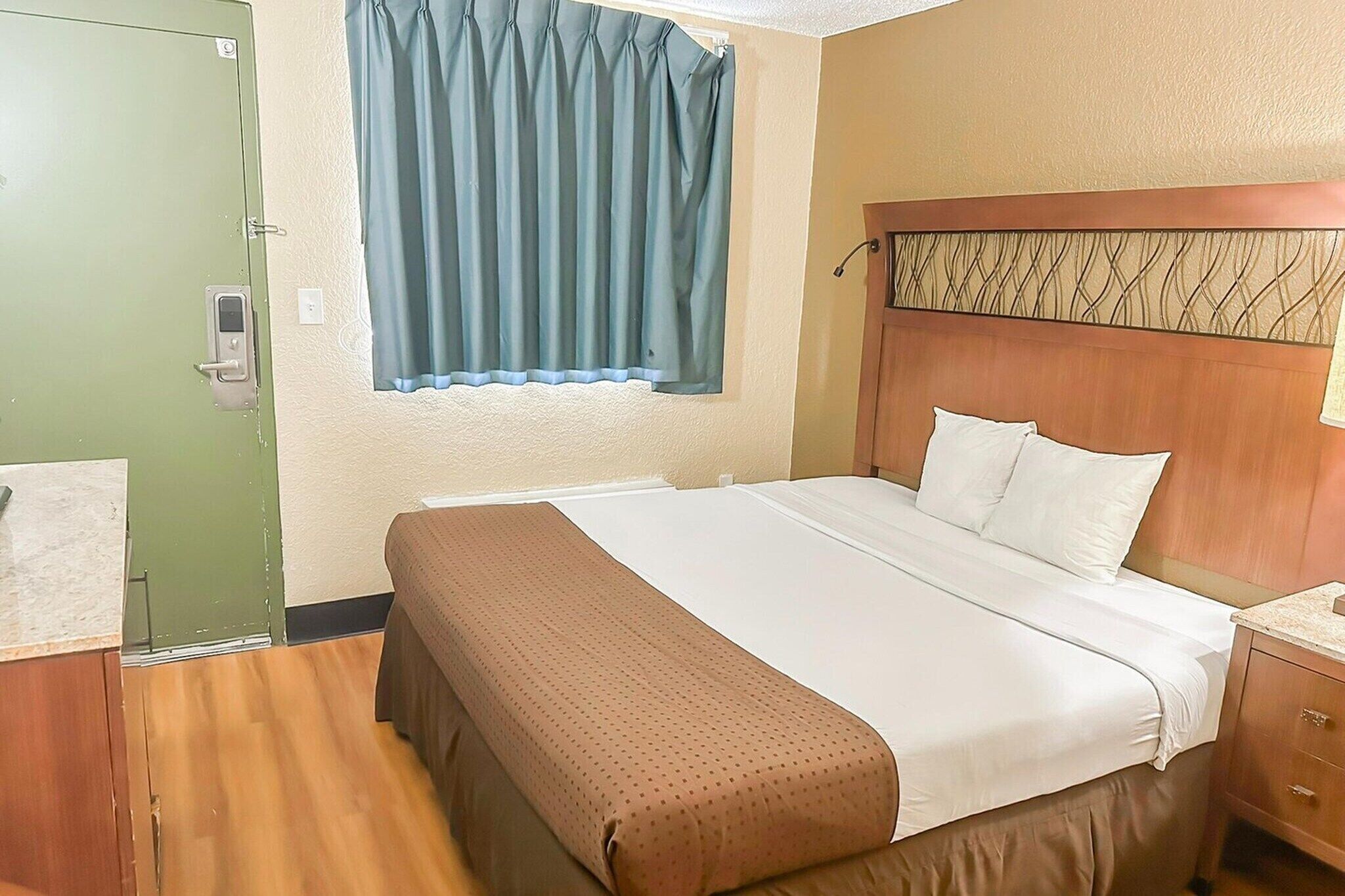 A hotel room with a bed, a wooden headboard, a window with curtains, a green door, and a wooden floor.