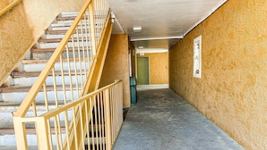 A narrow hallway with a staircase on the left, a green door at the end, and a trash bin in the center.