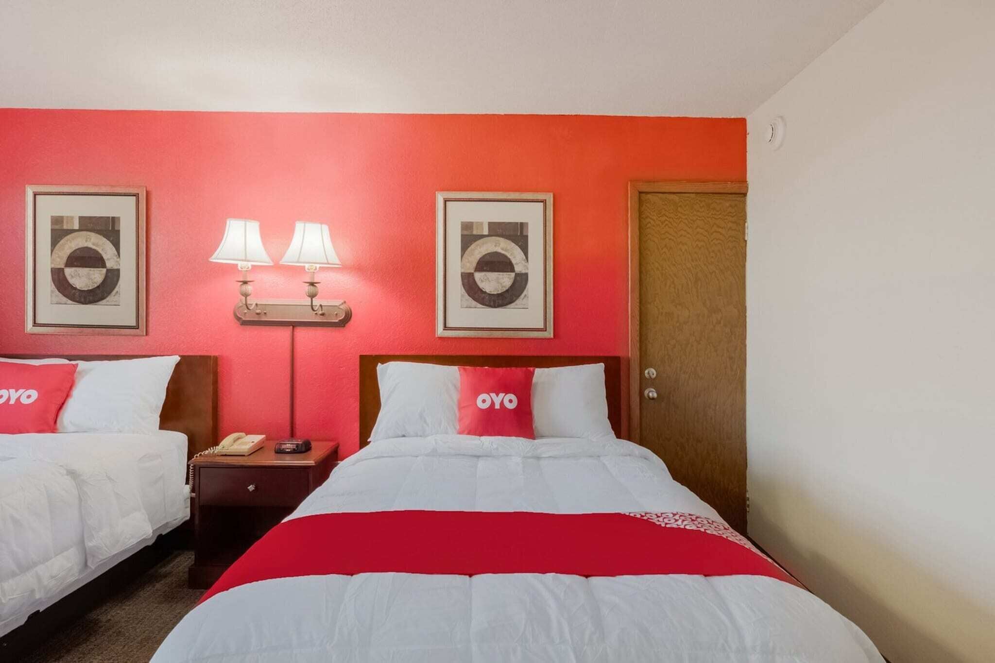 A hotel room with two beds, a red wall, a wooden door, and two framed pictures.