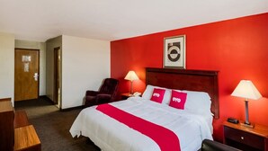 A hotel room with a red wall, a bed with white bedding and red pillows, two bedside lamps, a wooden headboard, a framed picture above the bed, and a wooden door. - King Bed Accessible|Nsmk (Morton IL)