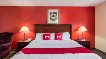 A hotel room with a red wall, a bed with white bedding and red pillows, two bedside lamps, a wooden headboard, and a framed picture above the bed.
