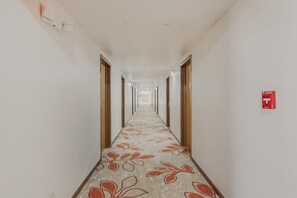 Un pasillo largo y estrecho con alfombra estampada, puertas de madera y un letrero de salida de emergencia rojo.